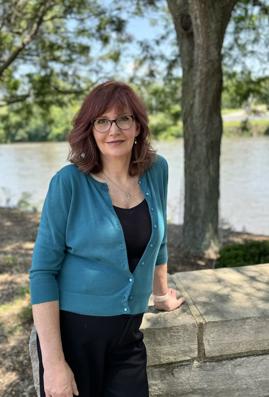 Woman smiling outdoors near river and trees.