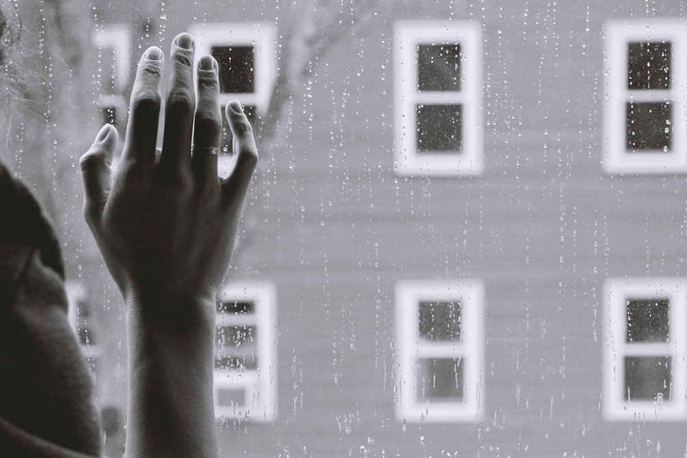 Hand reaching against rainy window, gray building outside.